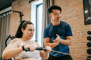 Low angle of smiling ethnic male trainer clapping hands to overweight female training on rowing machine in gym